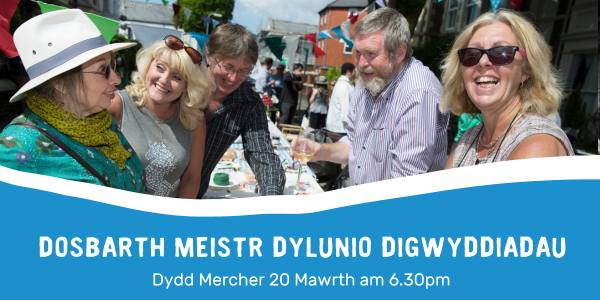 Group of residents smiling at Big Lunch table in a street with text: 'Dosbarth meistr dylunio digwyddiadau'