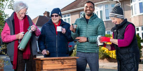 Four neighbours smiling and dribing tea around a planter on their street