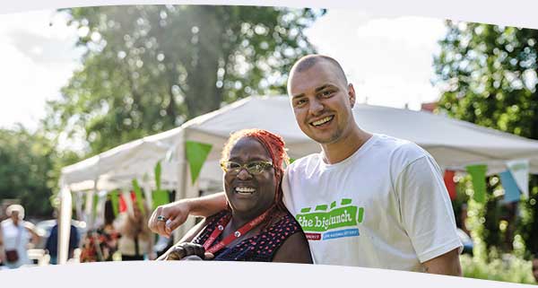 Young man and lady arm in arm smiling at a Big Lunch event outside with bunting and gazeebo in the background