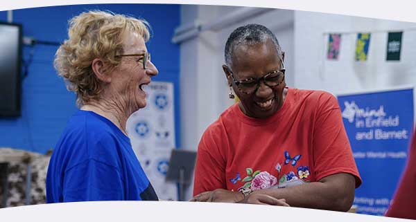 Two women laughing at a community event with Mind charity banner and Eden Project bunting in background